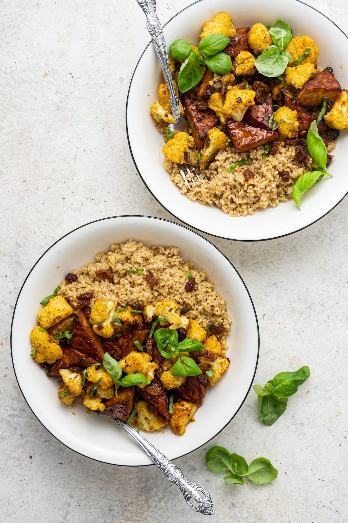 Two white bowls with metal forks containing the curried cauliflower and tempeh over a bed of quinoa, garnished with fresh basil leaves.