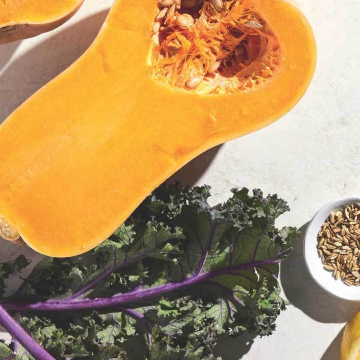A split butternut squash near a kale leaf and a small bowl of seeds.