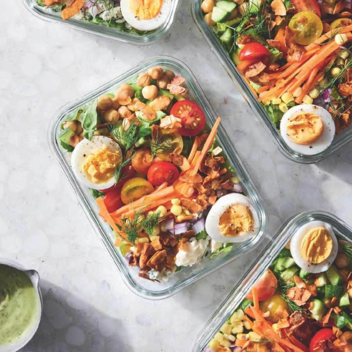 Four glass meal prep containers with hard boiled eggs, cherry tomatoes, carrots, beans, and fresh greens.