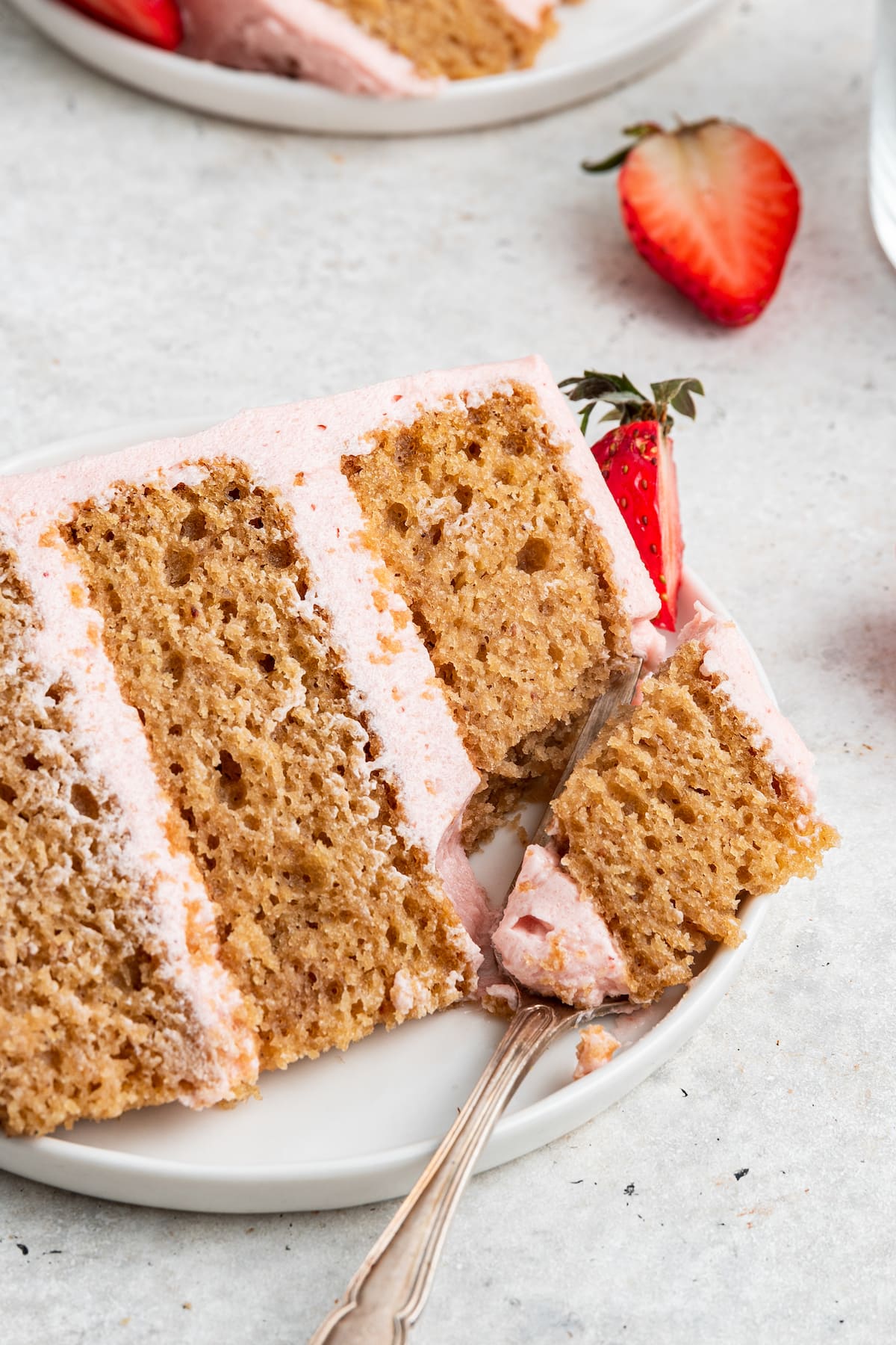 A slice of strawberry cake on a small white plate with a metal fork holding a piece of the slice.
