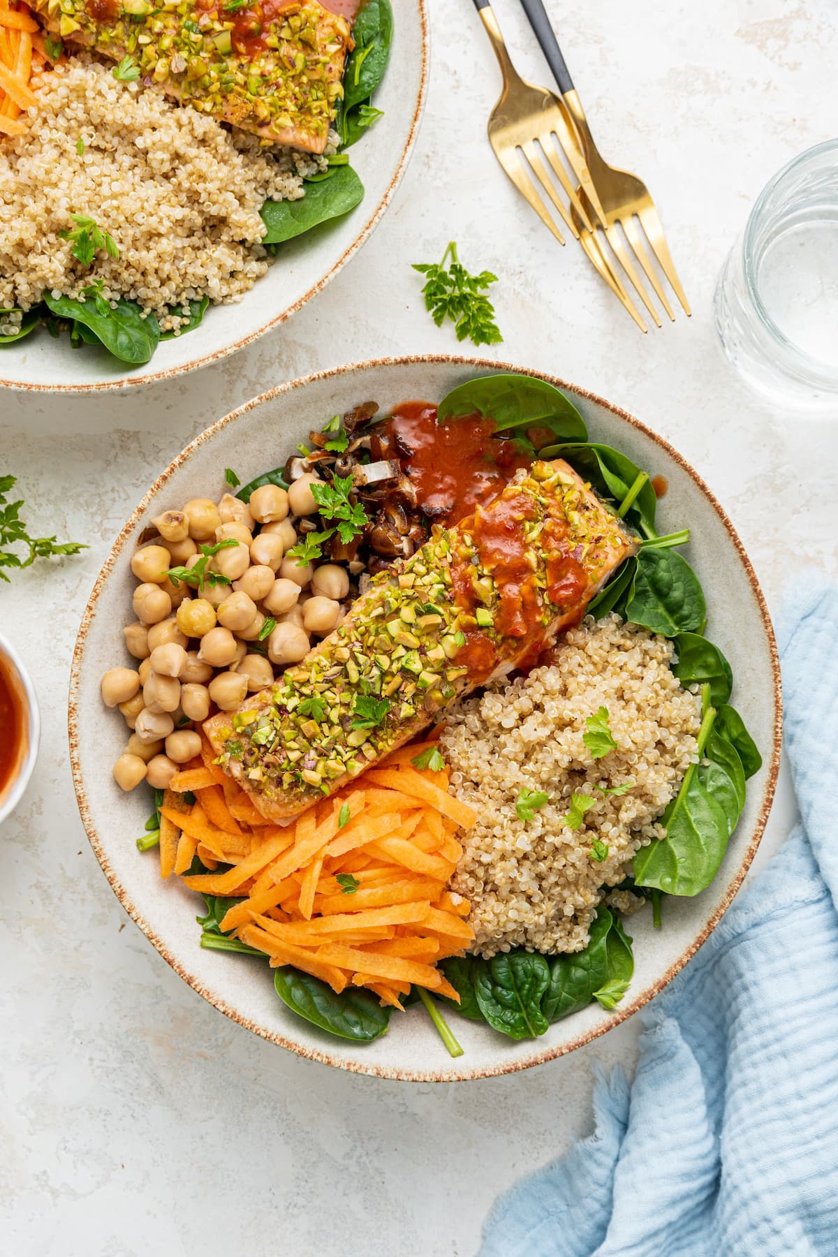A pistachio salmon salad served in a white bowl. The bowl contains a baked salmon filet, chickpeas, shredded carrots, quinoa, chopped dates, spinach, and spicy harissa dressing.