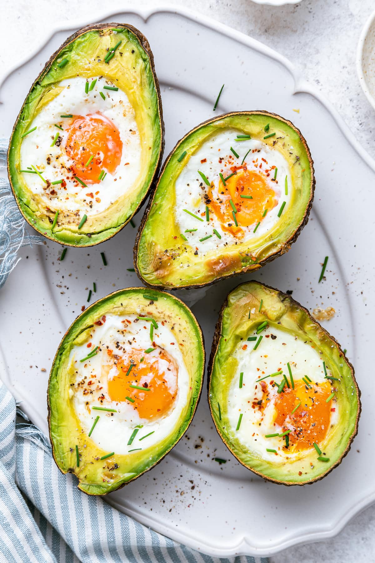 Four avocado halves with baked eggs in the center of each, served on a plate with fresh chives and paprika.