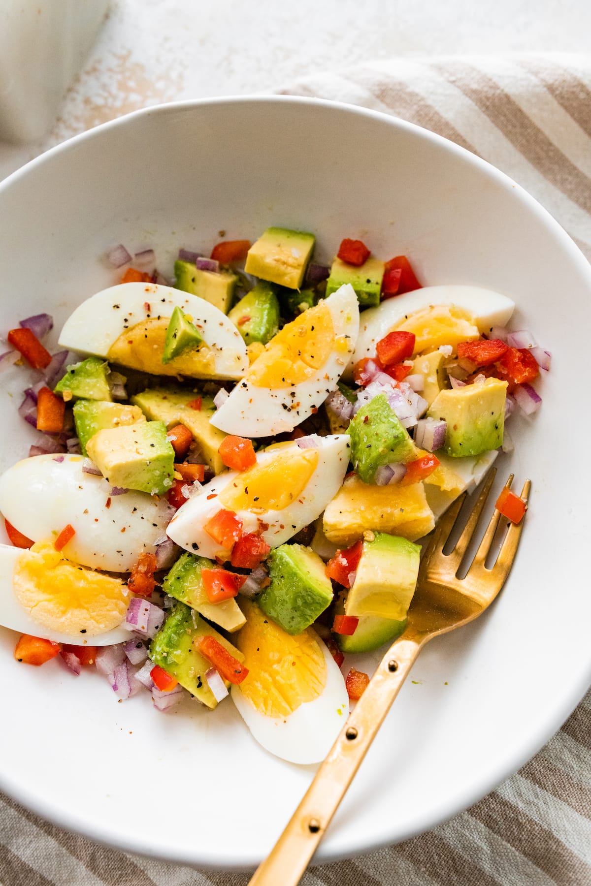 A serving of hard boiled eggs, diced avocado, red bell pepper, and onion in a bowl with a metal fork.