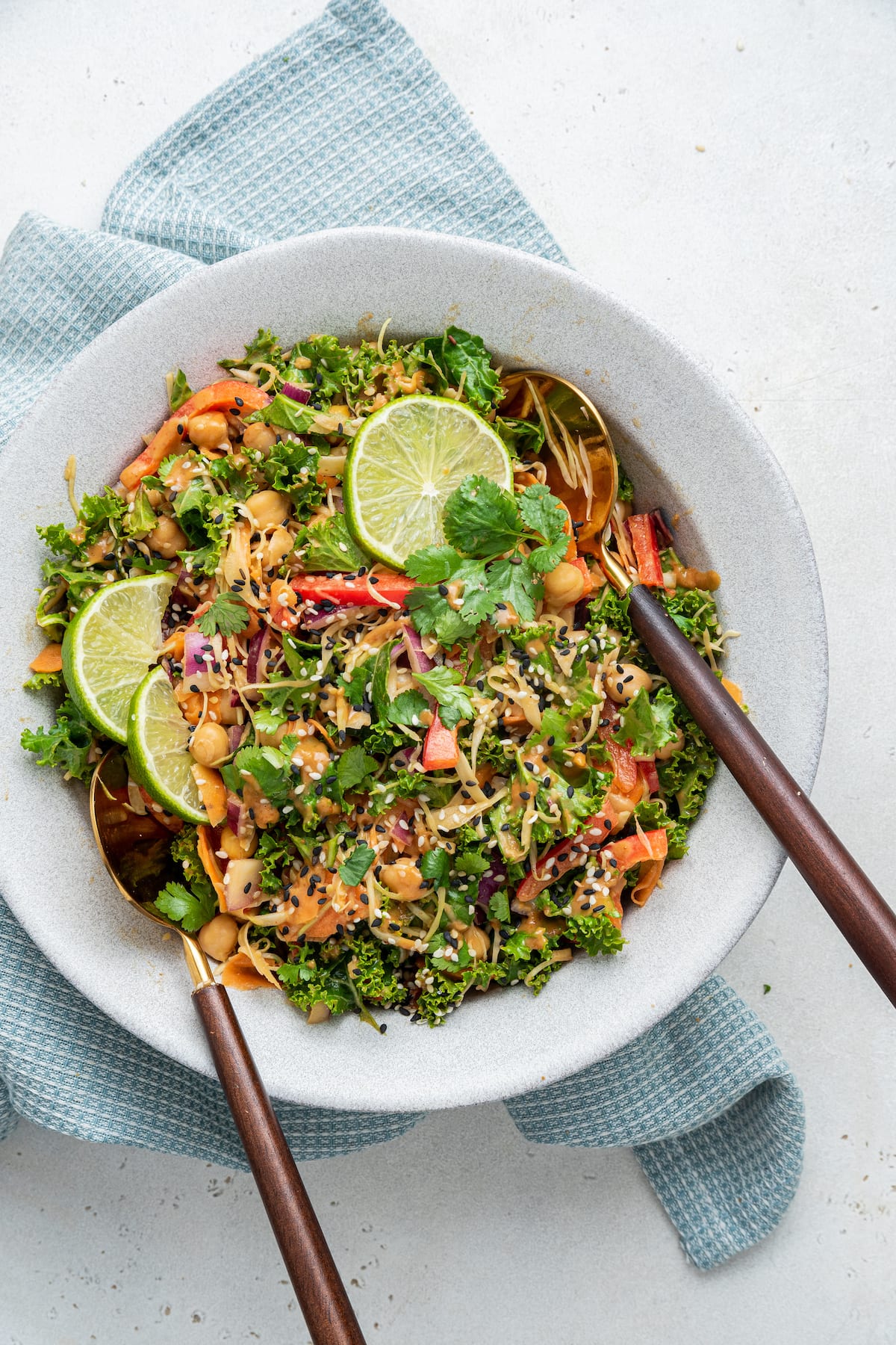 A bowl with a kale and pad Thai salad, two serving spoons, and garnished with a few lemon wedges.