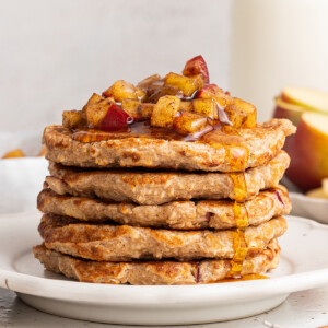 Five apple pancakes stacked on one another on a white plate with diced apples and maple syrup on top.
