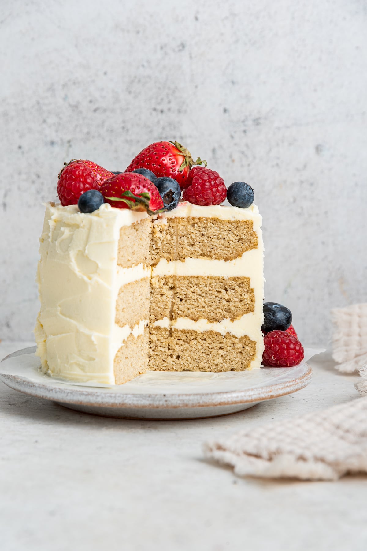 A frosted smash cake topped with fresh berries sitting on a plate. Once slice of the cake has been cut out of it.