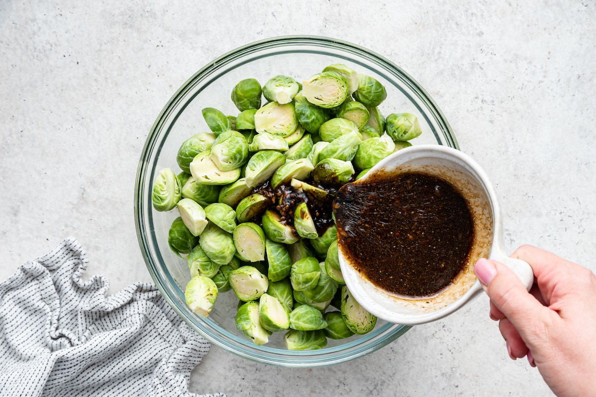 A woman's hand pouring a balsamic mixture over halved Brussels sprouts in a large glass bowl