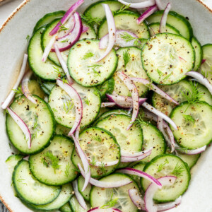 A large bowl with sliced cucumber, sliced red onion, dill, pepper, and a vinaigrette.
