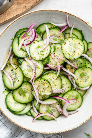 A large bowl with sliced cucumber, sliced red onion, dill, pepper, and a vinaigrette.