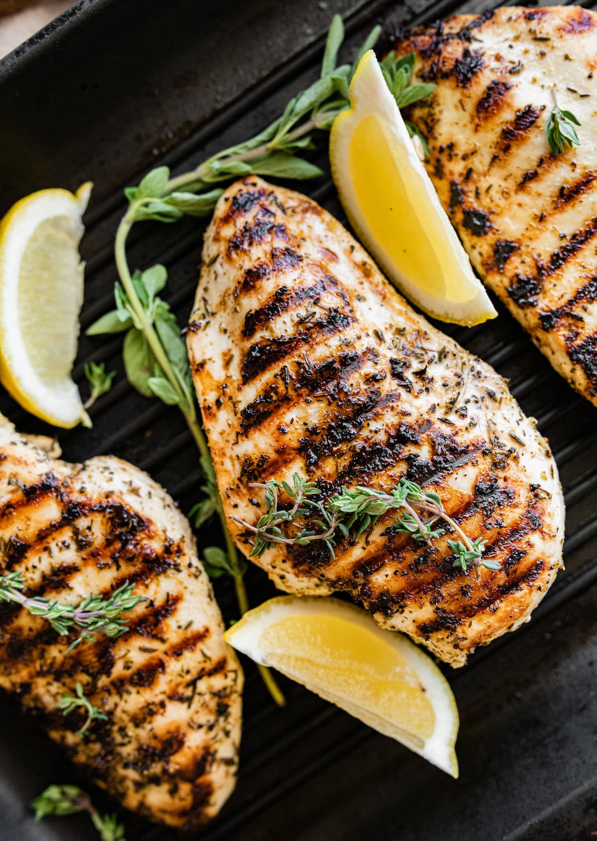 Grilled chicken breasts on a grill pan with fresh herbs and three lemon wedges nearby.