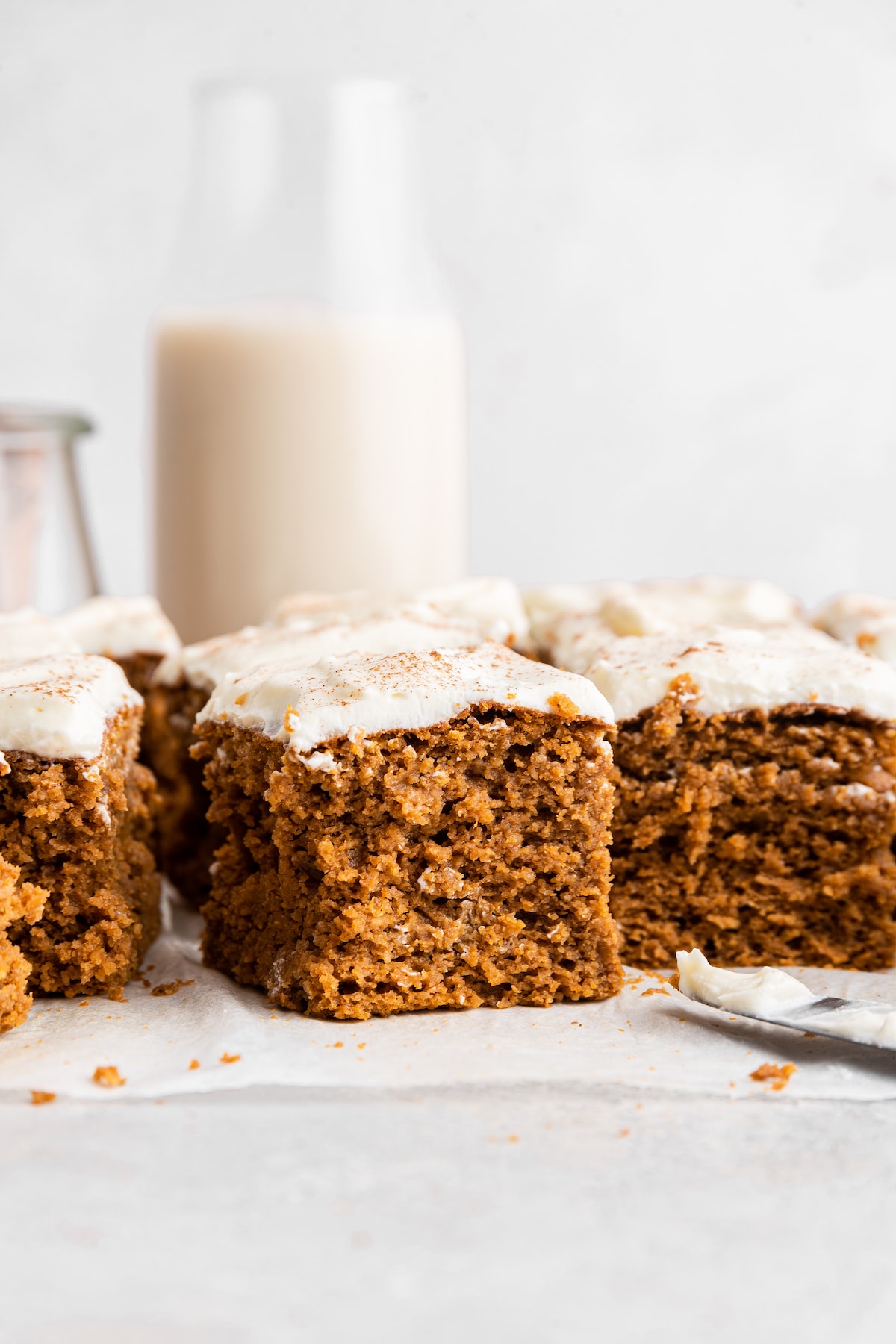 Multiple slices of pumpkin cake topped with a creamy vanilla frosting near a glass jar of milk.