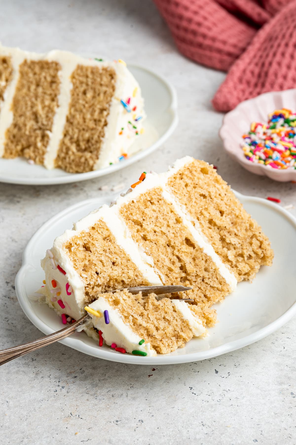 A slice of vanilla cake on a plate with a fork cutting a bite size piece from it.