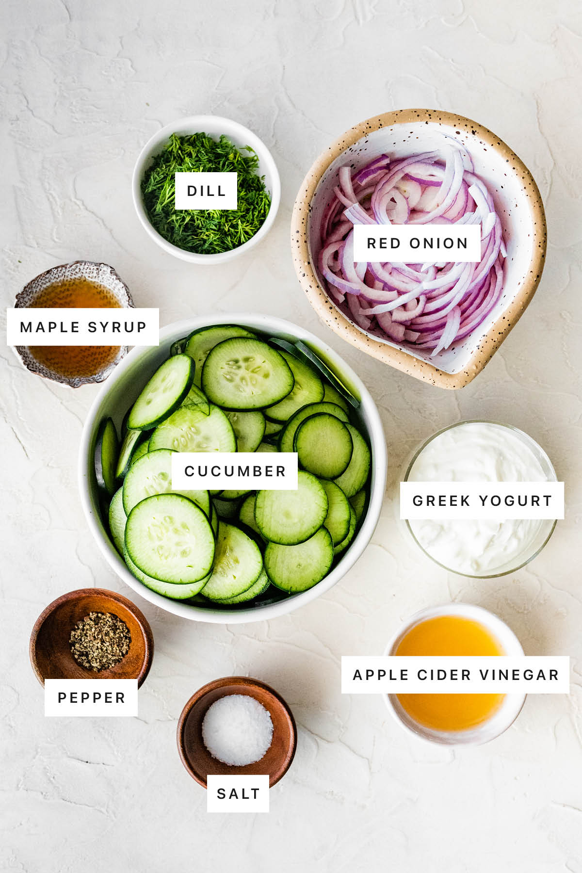 Ingredients measured out to make Creamy Cucumber Salad: dill, red onion, maple syrup, cucumber, Greek yogurt, pepper, salt and vinegar.