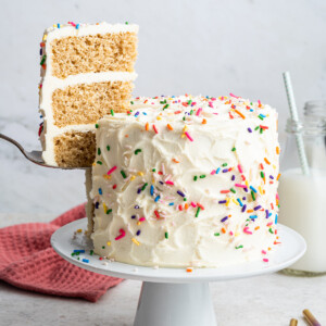A slice of cake is removed from the full vanilla cake that sits on a white pedestal. Cake is decorated with vanilla frosting and rainbow sprinkles.