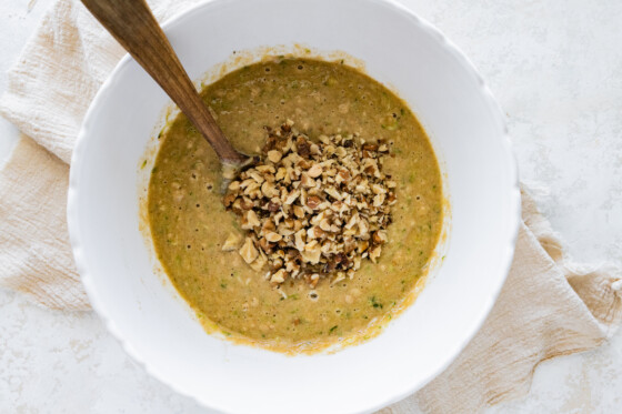 The batter for the healthy zucchini bread in a large mixing bowl with a wooden spoon and walnuts.