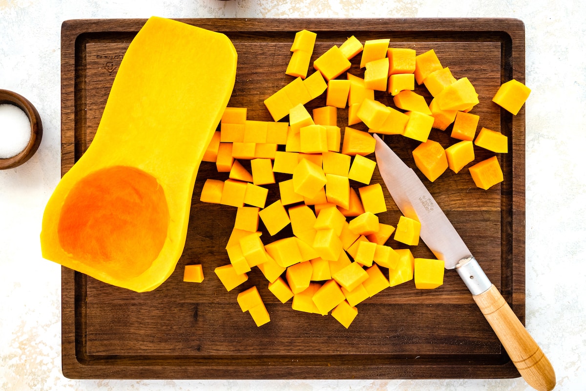 A deseeded butternut squash that has been cut in half on a wooden cutting board. One-half of the butternut squash has been cut into small cubes.