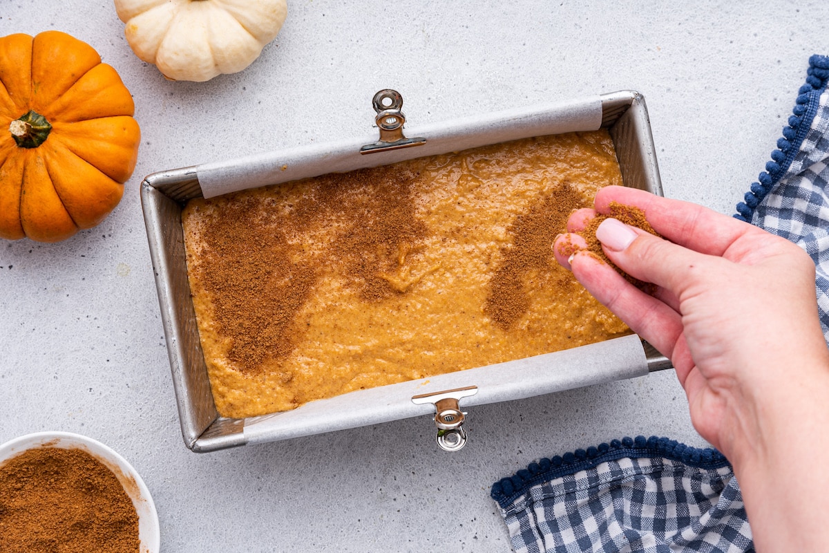 A woman's hand sprinkles a mixture of ground cinnamon and coconut sugar on top of the batter for healthy pumpkin bread in a loaf pan.
