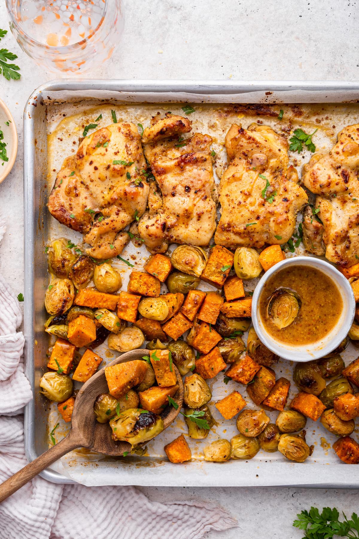 A sheet pan with honey mustard chicken, roasted sweet potato, brussels sprouts, and a wooden serving spoon.