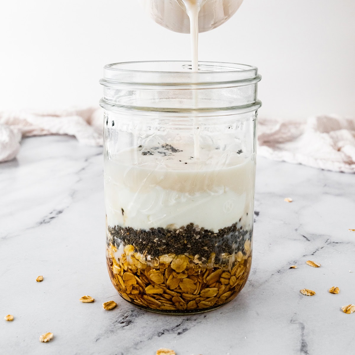 Oats, chia seeds, maple syrup and yogurt layered in a mason jar for overnight oats with milk being poured on top.