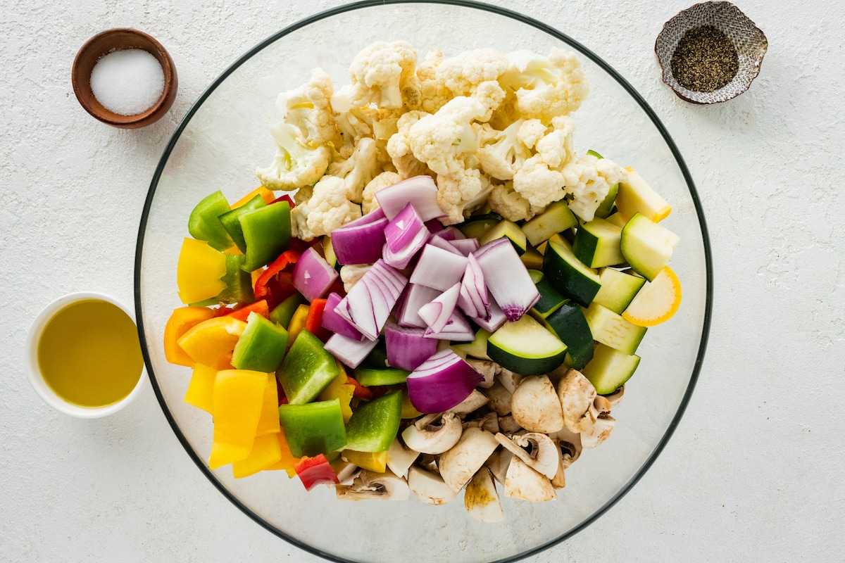 Raw vegetables in a large glass bowl. Vegetables include cauliflower, onion, zucchini, mushroom, and bell peppers.