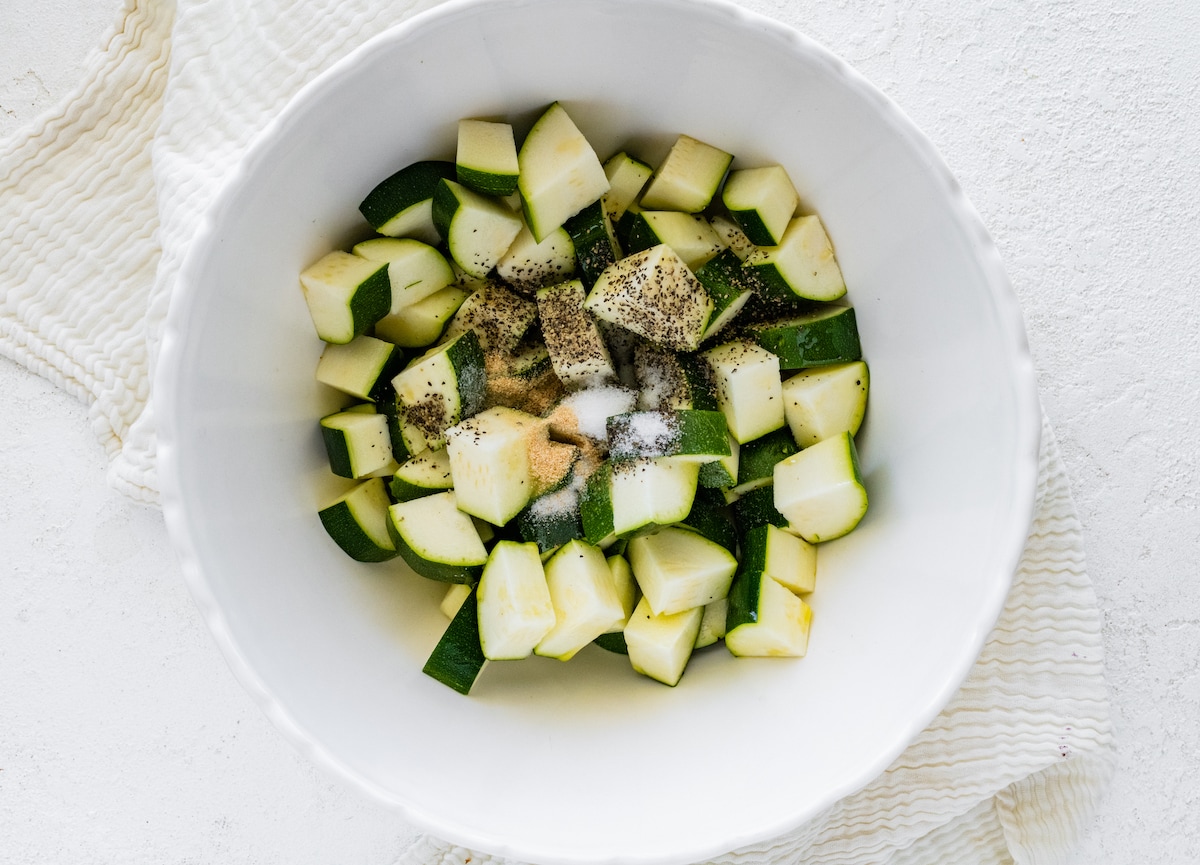 Chopped zucchini in a white bowl with seasonings added.