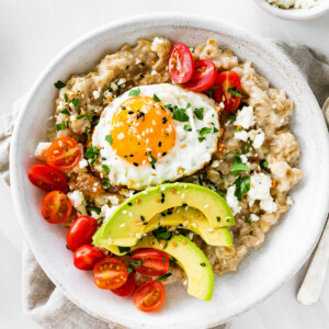 A serving of oatmeal in a bowl topped with cherry tomatoes, feta cheese, avocado slices, chives, salt, pepper, and a sunny side up cooked egg.