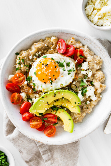 A serving of oatmeal in a bowl topped with cherry tomatoes, feta cheese, avocado slices, chives, salt, pepper, and a sunny side up cooked egg.