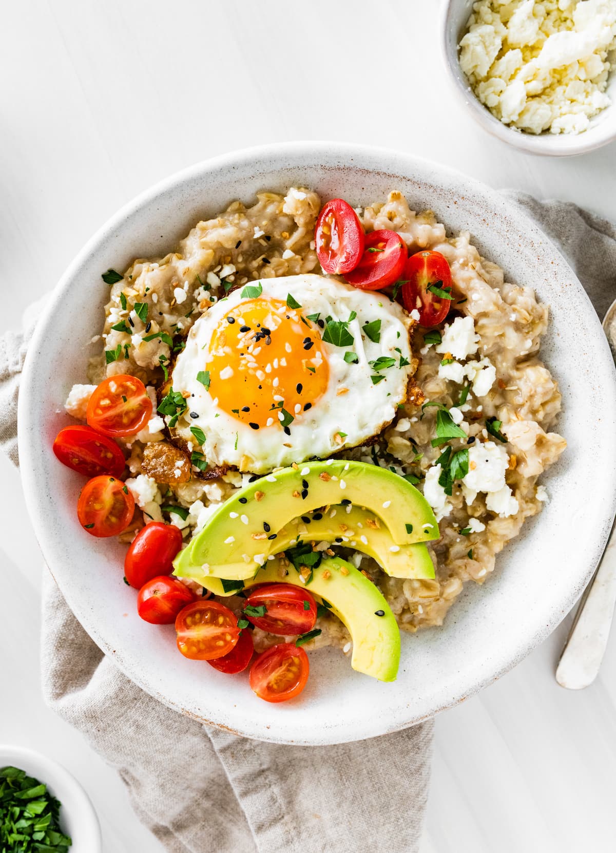 A serving of oatmeal in a bowl topped with cherry tomatoes, feta cheese, avocado slices, chives, salt, pepper, and a sunny side up cooked egg.