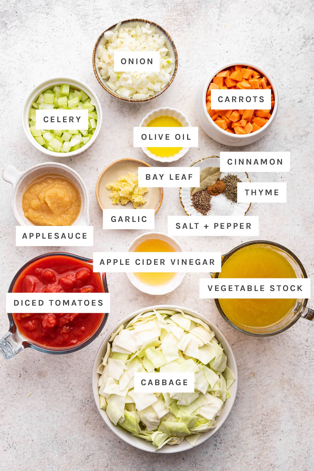 Ingredients measured out to make cabbage soup: applesauce, garlic, salt, pepper, bay leaf, thyme, diced tomatoes, onion, olive oil, cinnamon, vegetable stock, cabbage, carrots, celery, and vinegar.