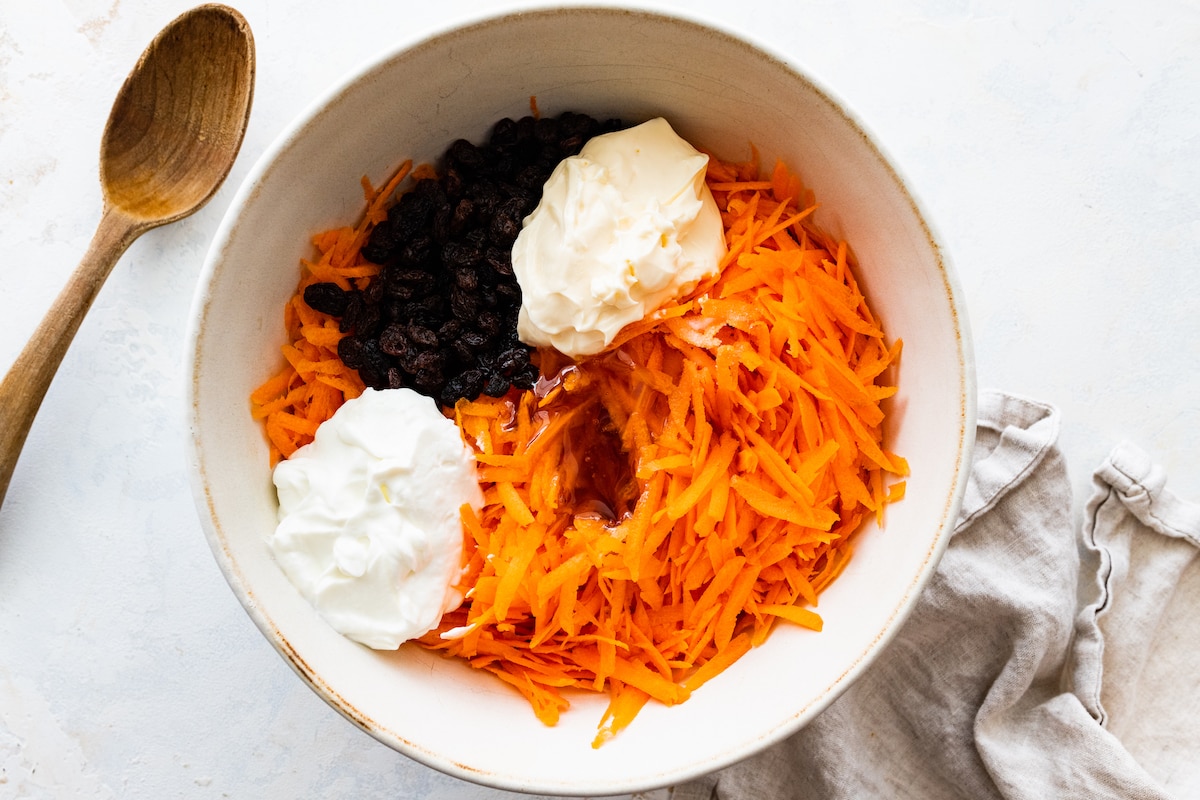 Grated carrots added to a large mixing bowl along with raisins and yogurt.
