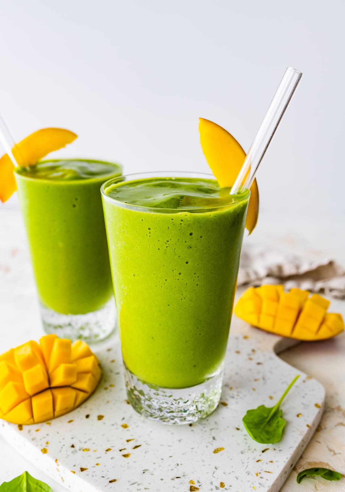 Two green smoothies in glasses with straws and fresh mango slices.