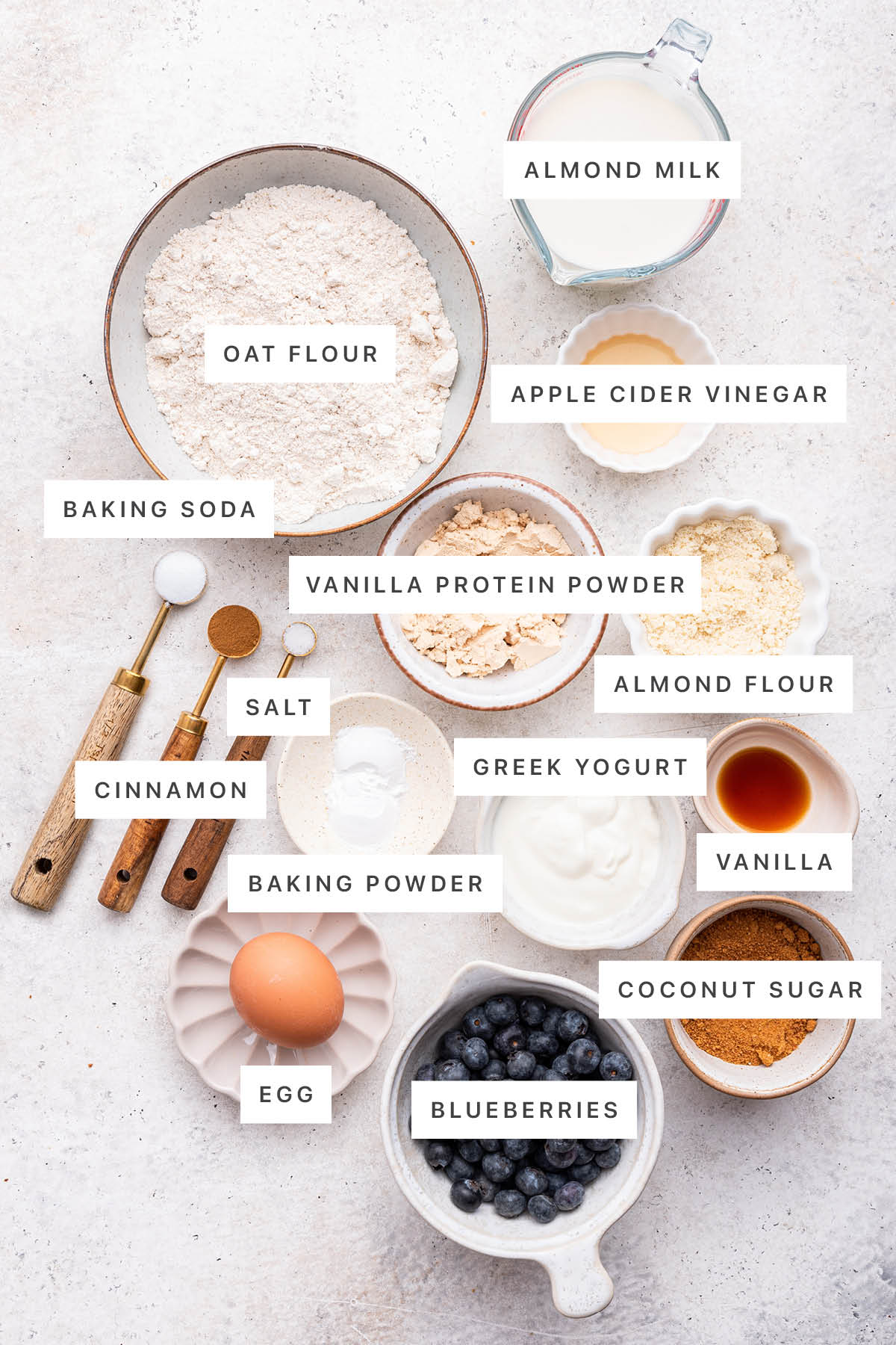 Ingredients measured out to make blueberry protein muffins: oat flour, almond milk, vinegar, baking soda, cinnamon, salt, vanilla protein powder, almond flour, greek yogurt, baking powder, vanilla, coconut sugar, an egg, and blueberries.