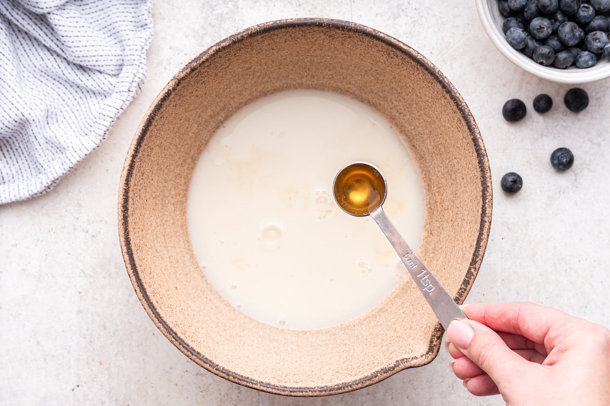 A woman's hand holding a teaspoon of vanilla over a large mixing bowl of milk.