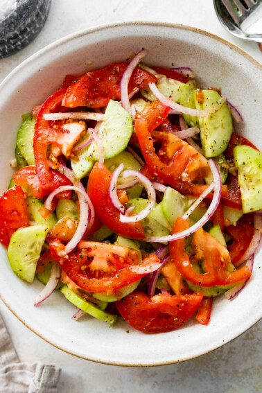In a small white bowl, a cucumber tomato salad with sliced red onion, salt, and pepper.