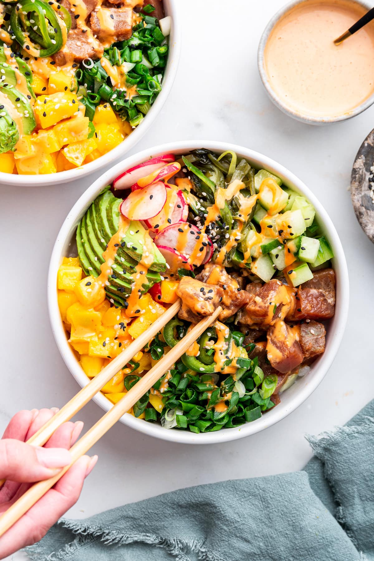 A woman's hand holds chopsticks picking up a piece of ahi tuna from a poke bowl. Poke bowl is topped with cucumbers, radishes, avocado, mango, scallions and jalapeños.