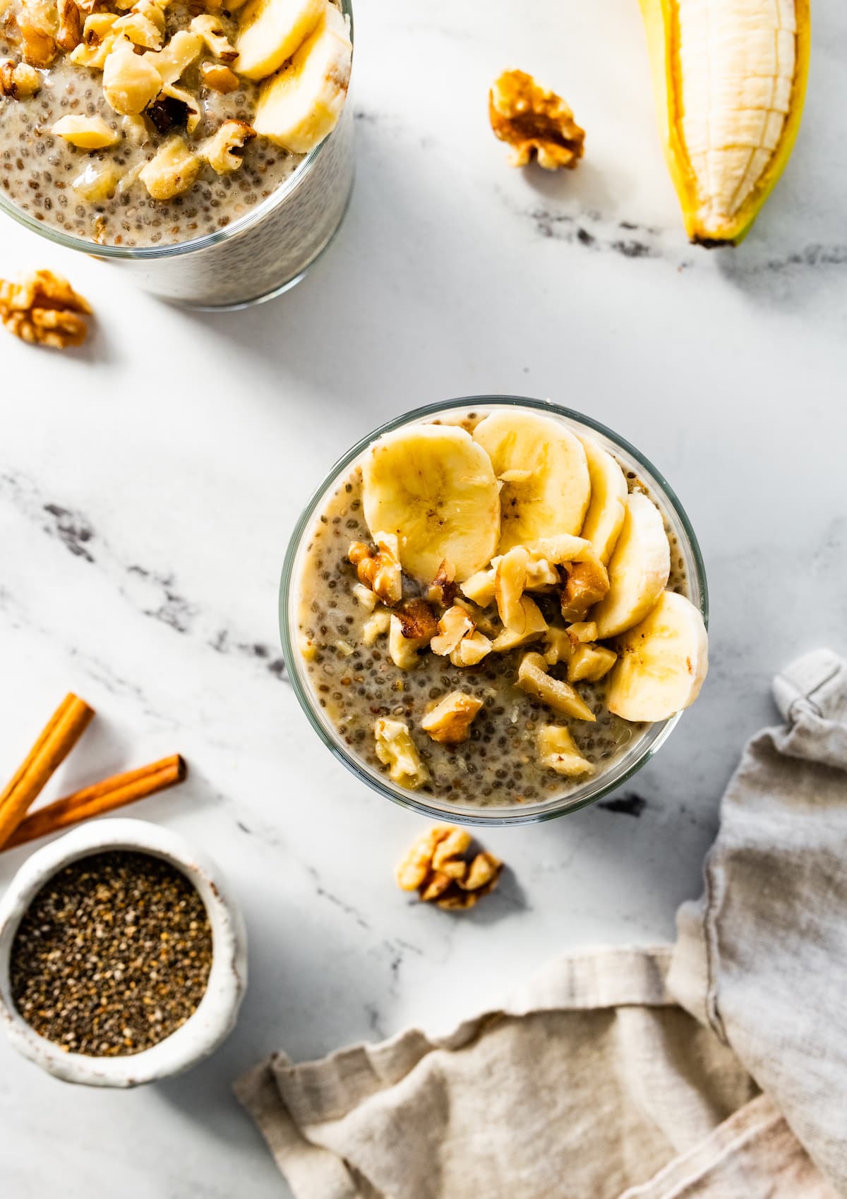 A serving of banana chia pudding in a glass cup topped with banana, and crushed walnuts.
