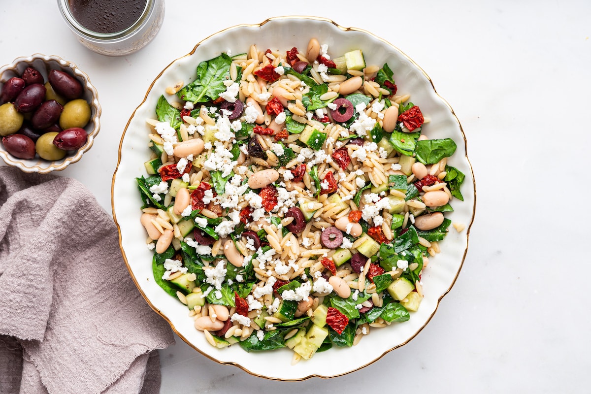 A large bowl with Mediterranean orzo salad with beans, spinach, sun dried tomatoes, diced cucumber, red onion, and olives.