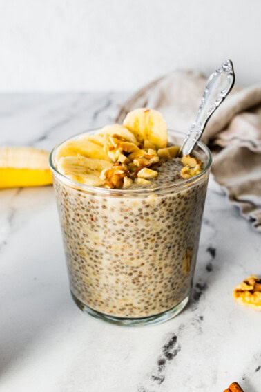 A glass cup with a serving of banana chia pudding that is topped with banana slices and walnuts.
