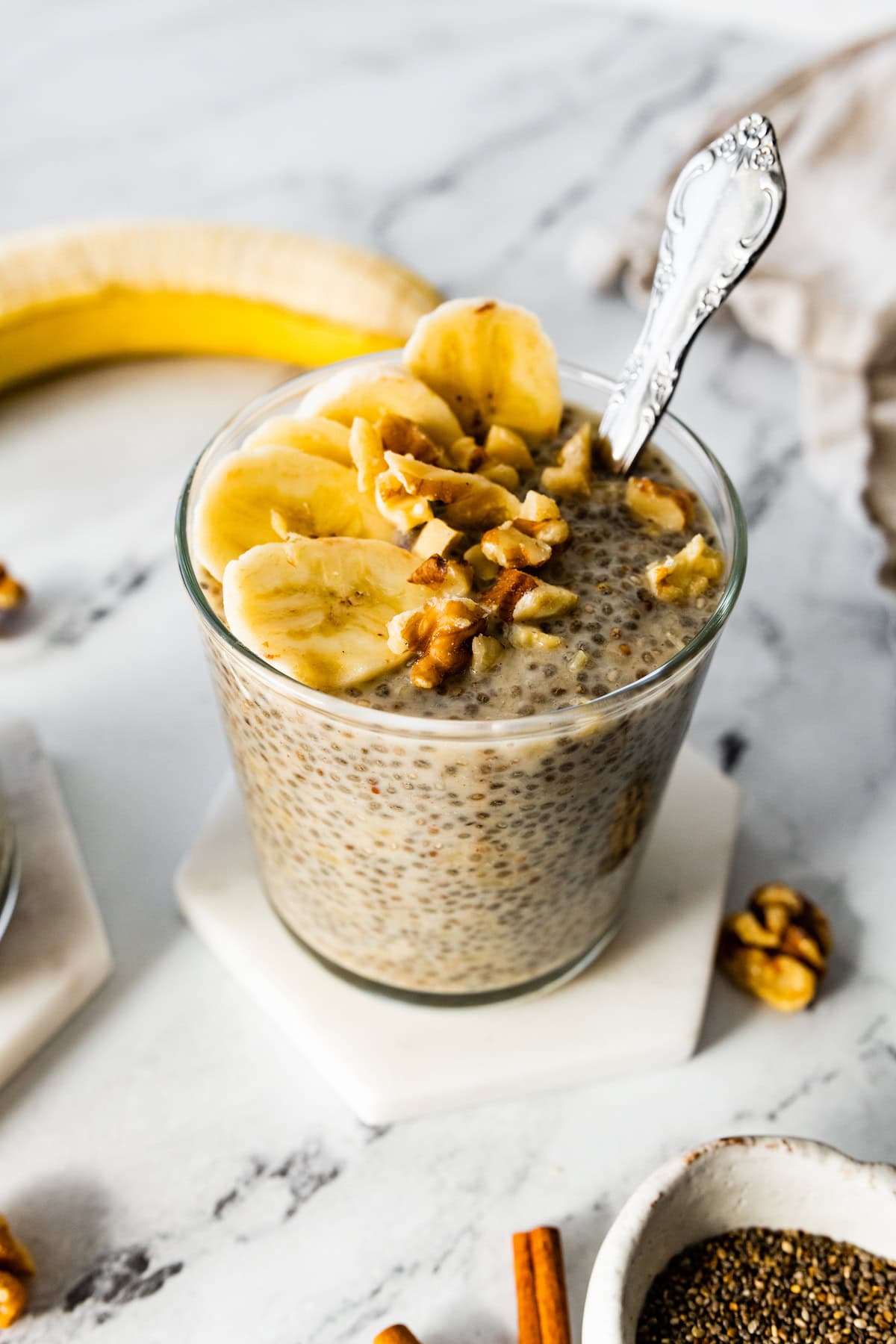 Banana chia pudding in a glass cup with a metal spoon and topped with banana slices and crushed walnuts.