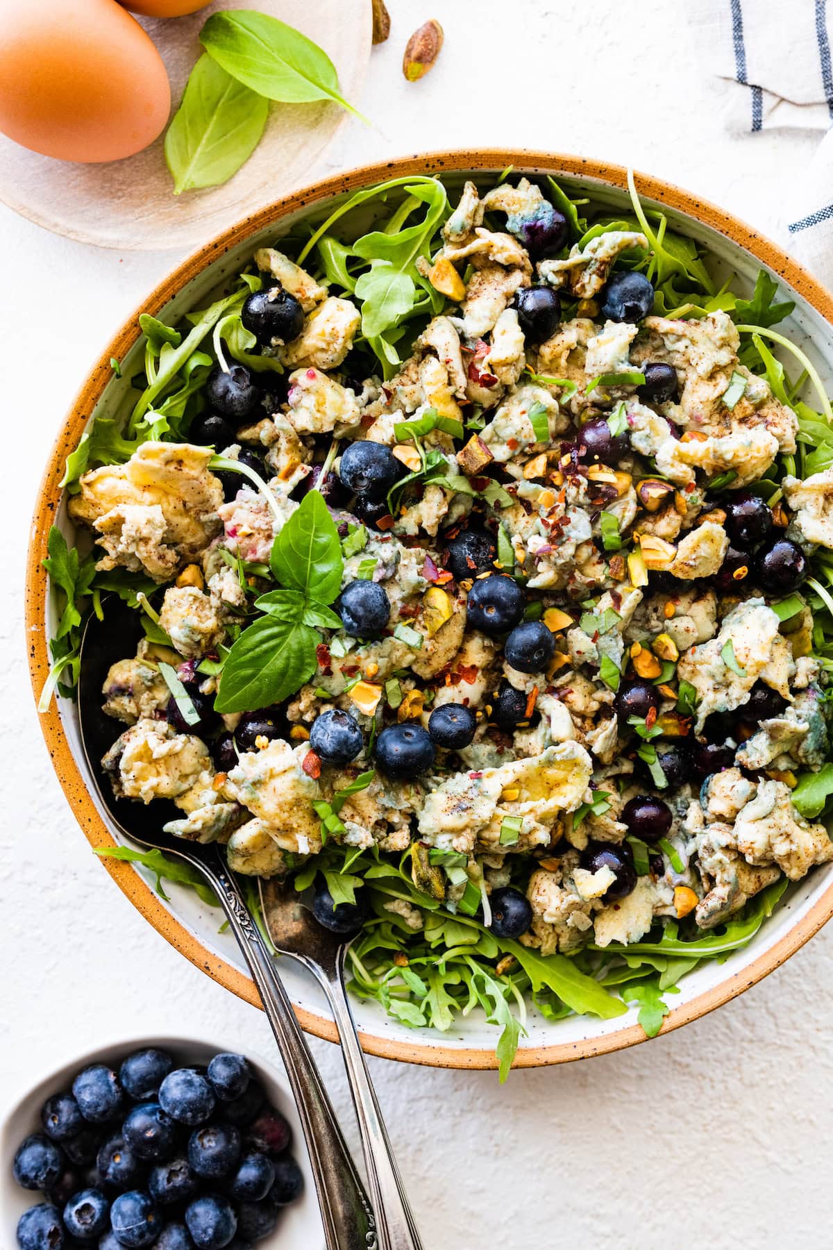 A large bowl with two metal spoons, scrambled eggs, arugula, blueberries, fresh basil leaves, herbs and spices.