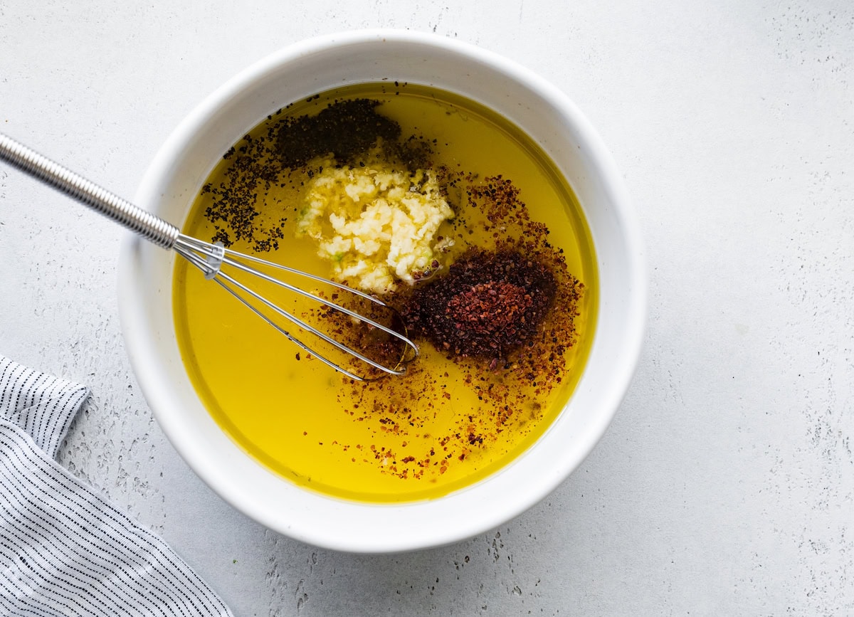 A metal whisk in a small bowl of minced garlic, olive oil, lemon, and spices.