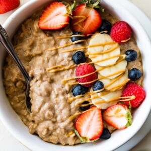 A serving of protein oatmeal topped with fresh blueberries, raspberries, strawberries, bananas, and a drizzle of peanut butter all in a white bowl.