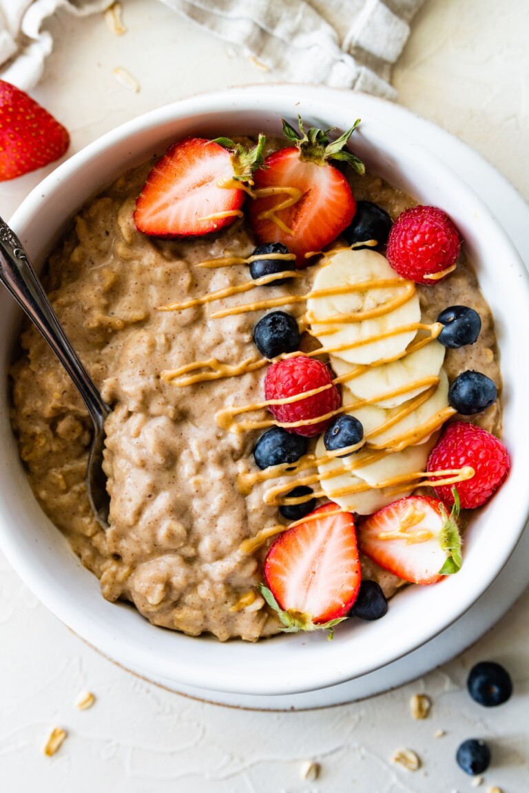 A serving of protein oatmeal topped with fresh blueberries, raspberries, strawberries, bananas, and a drizzle of peanut butter all in a white bowl.