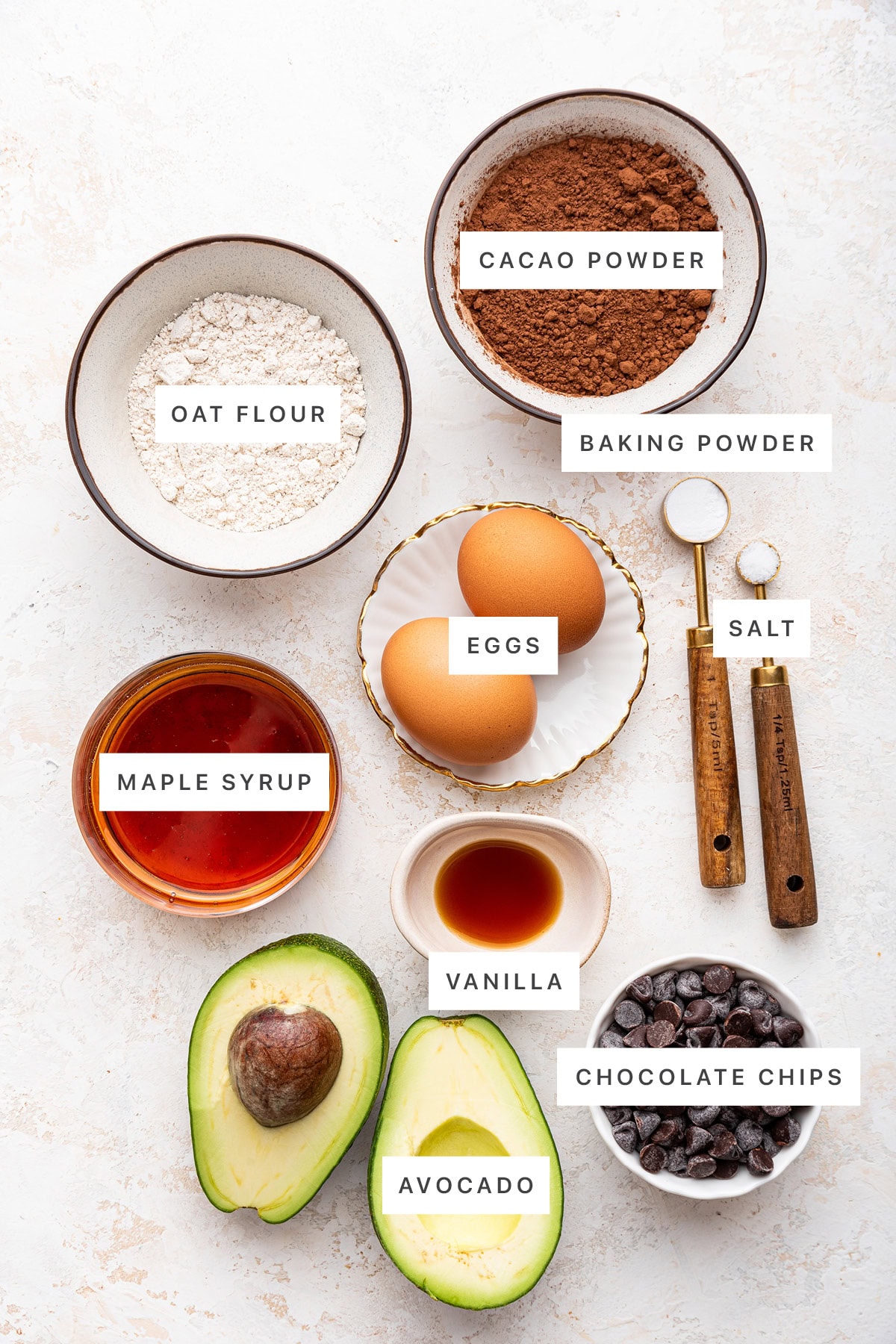 Ingredients measured out to make Avocado Brownies: oat flour, cacao powder, baking powder, salt, eggs, maple syrup, vanilla, avocado and chocolate chips.
