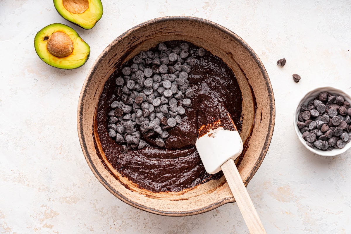 A large mixing bowl of avocado brownie batter with chocolate chips and a silicone spatula.