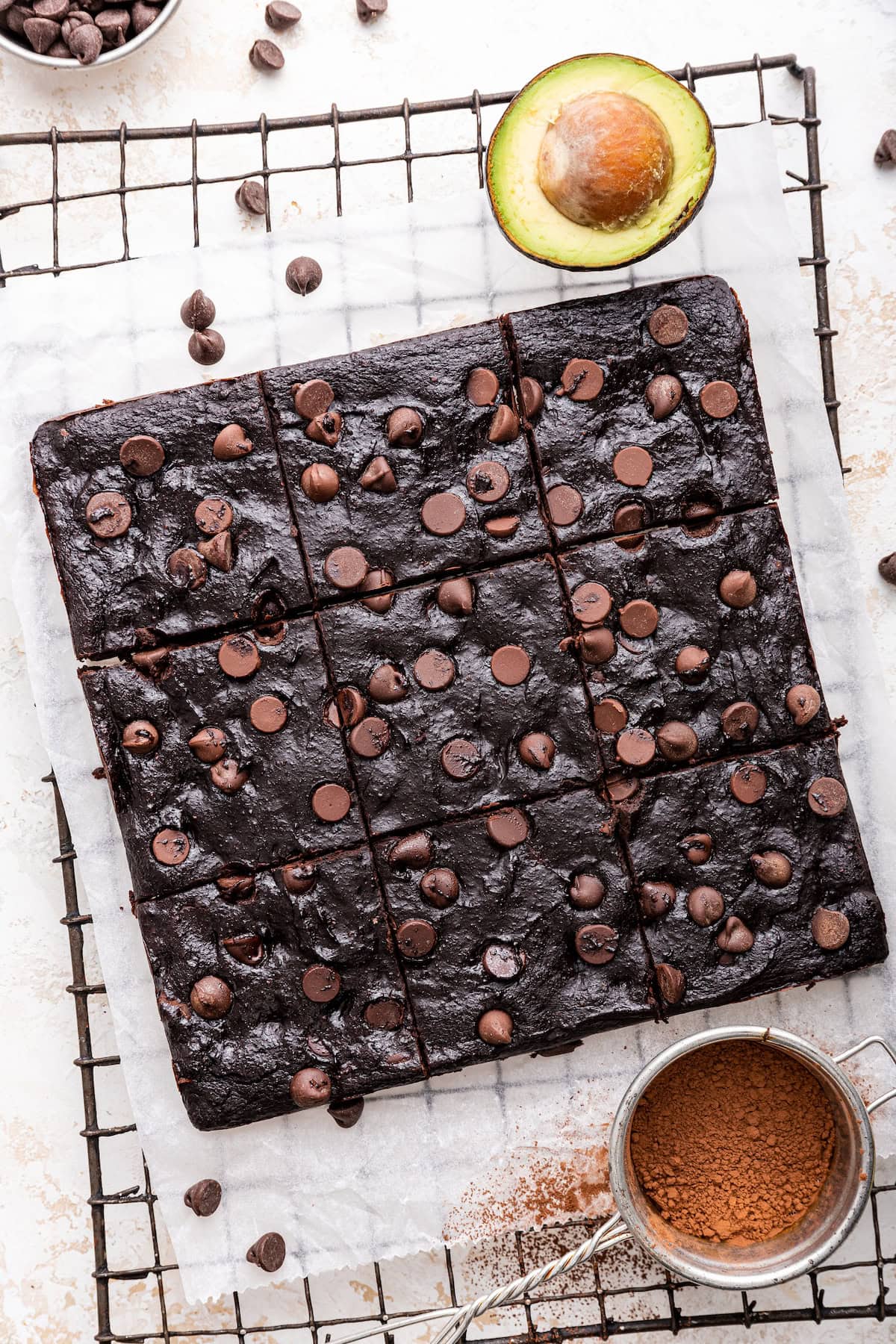 Cut chocolate chip avocado brownies on a wire rack. There is an avocado and a sifter with coco powder near the brownies.
