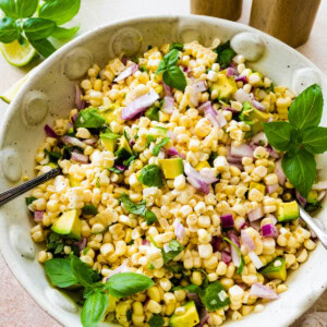 A large bowl with corn, diced avocado, red onion, fresh basil, and two serving spoons.