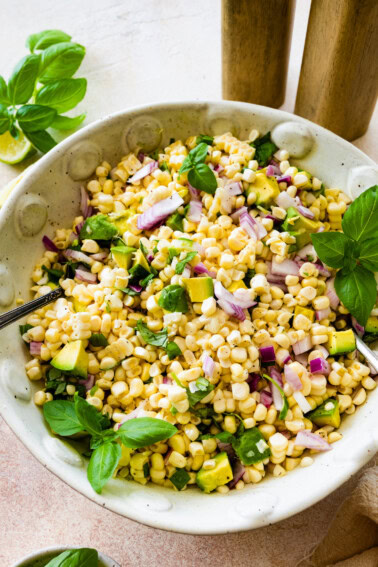 A large bowl with corn, diced avocado, red onion, fresh basil, and two serving spoons.
