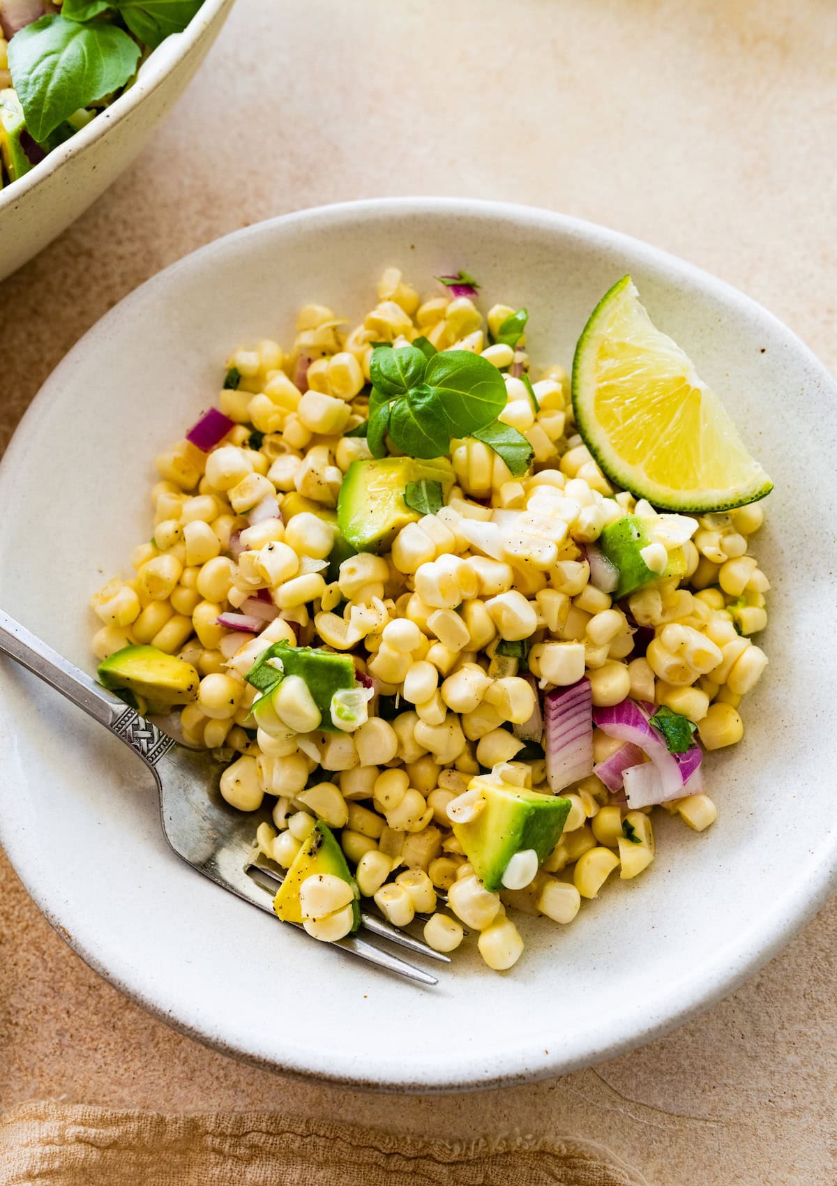 A serving of avocado corn salad on a small plate with a metal fork and a lime wedge on the side of the plate.