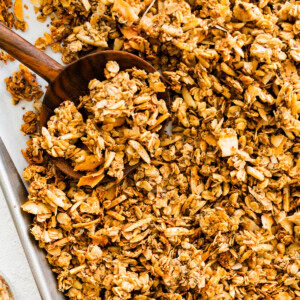 A baking sheet with a large wooden spoon taking a portion of coconut granola.